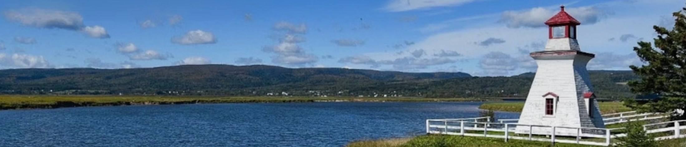 A white lighthouse overlooking the water and green hills of Albert County